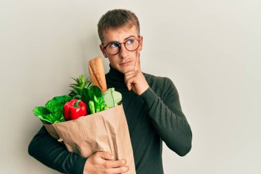 Elinde alışveriş poşeti olan beyaz bir genç adam ciddi bir yüzle eli çenesinde soru sormayı düşünüyor. Kafa karıştırıcı bir fikir. 