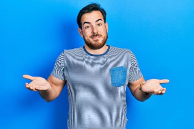 Sakallı, sıradan çizgili tişörtsüz, kafası karışmış, elleri ve kolları havada yakışıklı beyaz bir adam. şüphe kavramı. 