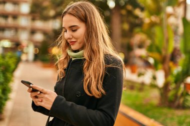 Genç sarışın kız şehirde akıllı telefon kullanarak mutlu bir şekilde gülümsüyor..