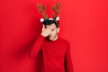 Şirin Noel geyiği boynuzları takan genç İspanyol adam şok içinde yüzünü ve gözlerini kapayarak utangaç ifadesiyle parmaklarından bakıyor.. 