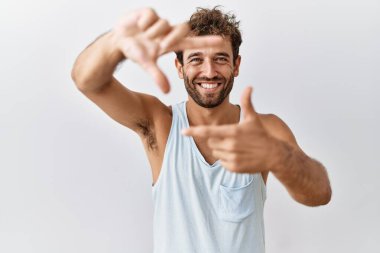 İzole edilmiş arka planda duran yakışıklı genç adam gülümsüyor ve elleri ve parmakları gülen yüzlü bir çerçeve yapıyor. Yaratıcılık ve fotoğrafçılık kavramı. 