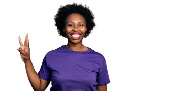 Afro saçlı, sıradan mor tişörtlü Afro-Amerikalı bir kadın üç numaralı parmaklarını gösterirken kendinden emin ve mutlu bir şekilde gülümsüyor.. 