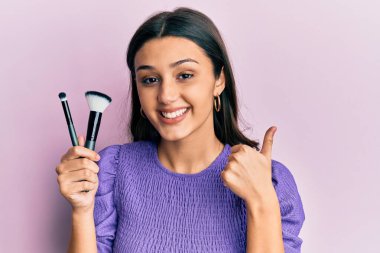 Makyaj fırçası tutan genç İspanyol kadın mutlu ve olumlu gülümsüyor, başparmağıyla mükemmel ve onay işareti yapıyor. 