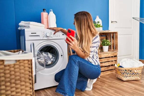 Çamaşır Odasında Akıllı Telefon Kullanan Çamaşır Yıkayan Genç Sarışın Kadın — Stok fotoğraf