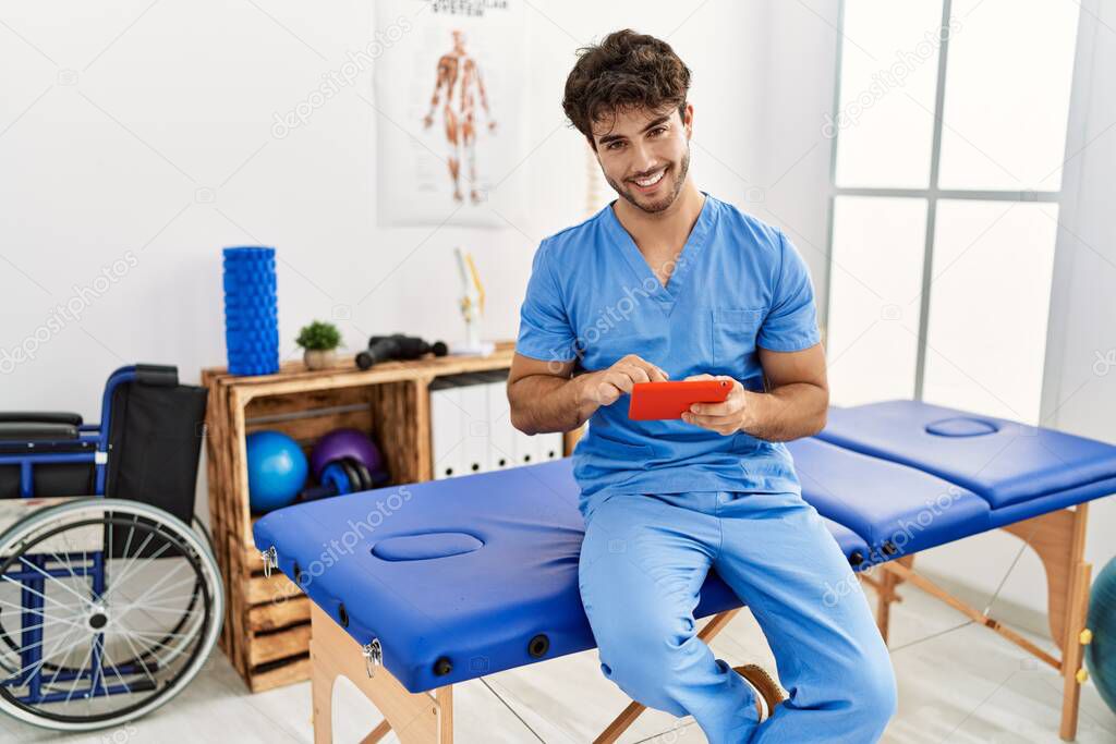 Young hispanic man wearing physio therapist uniform using touchpad at clinic