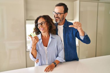 Orta yaşlı çift, mutlu bir şekilde gülümseyerek yan tarafa bakıyor ve başparmağıyla işaret ediyor.. 