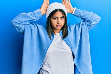 Şirin yün şapkalı, tavşan kulağı jestleri yapan, avuçlarıyla alaycı ve şüpheci görünen genç Latin bir kadın. Paskalya tavşanı konsepti. 