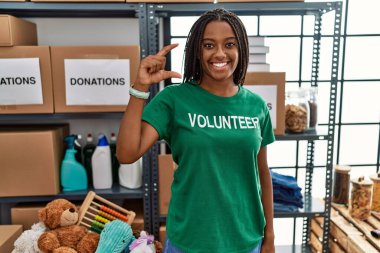 Bağışlarda gönüllü tişört giyen Afro-Amerikalı genç bir kadın gülümsüyor ve el hareketleriyle küçük boy işareti yaparak kameraya bakıyor. ölçüm kavramı. 