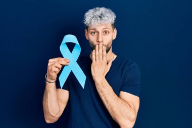 Modern boyalı saçları olan genç İspanyol bir adam elinde pembe kanser kurdelesiyle ağzını eliyle kapatıyor, şok geçiriyor ve hata yapmaktan korkuyor. şaşırmış ifade 