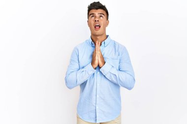 İspanyol iş gömleği giyen genç adam izole edilmiş arka planda duruyor. Elleriyle yalvarıyor ve dua ediyor. Yüzünde umut ifadesi var. Yalvarıyor. 