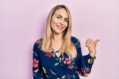 Gündelik kıyafetler giyen, mutlu yüzlü gülümseyen ve baş parmağıyla yan tarafı gösteren beyaz bir kadın.. 