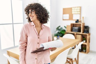 Ofiste iş elbisesi giyen genç bir ortadoğulu kadın yüzünde gülümseme, doğal bir ifade ile yan yana bakıyor. Kendinden emin gülen. 