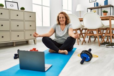 Orta yaşlı beyaz bir kadın evde laptopla yoga eğitimi veriyor.