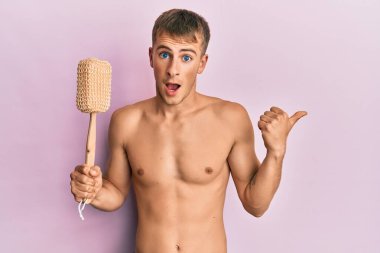 Beyaz, üstsüz, duş süngeri tutan genç bir adam başparmağıyla yan tarafa bakıyor ve ağzı açık gülümsüyor. 