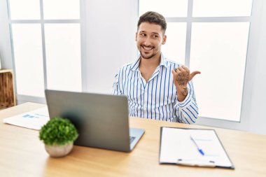 Ofiste çalışan sakallı, mutlu yüzüyle gülümseyen ve baş parmağıyla yüzünü gösteren bilgisayar bilgisayarı kullanan genç yakışıklı bir adam.. 