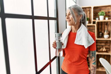 Orta yaşlı, gri saçlı, gülümseyen, elinde bir şişe su tutan bir kadın.