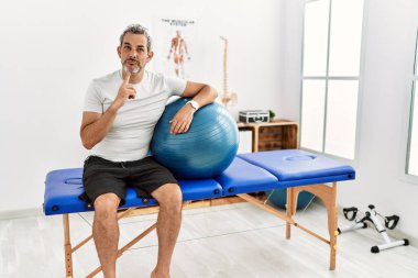 Orta yaşlı İspanyol bir adam ağrıyı dindirme kliniğinde elinde pilates topu tutarken parmakları çenesinde şüpheye yoğunlaşarak yukarı bakıyor. 