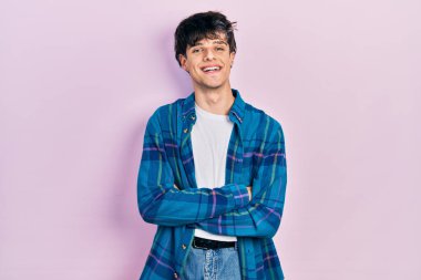 Yakışıklı, beyaz tişört ve klasik tişört giyen, kollarını kavuşturup kameraya gülümseyen genç bir hippi. pozitif kişi. 