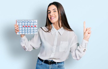 Elinde kalp takvimi olan genç esmer bir kız bir fikir veya soruyla gülümsüyor ve mutlu bir yüzle işaret ediyor. 