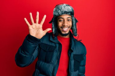 Sakallı, kışlık şapkalı ve paltolu genç Afrikalı Amerikalı beş numaralı parmaklarıyla işaret ederken kendinden emin ve mutlu bir şekilde gülümsüyor.. 