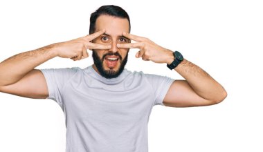 Sakallı, günlük beyaz tişörtlü genç adam barış sembolü yapıyor. Yüzünde parmaklar, gülümseyerek zaferi gösteriyor. 