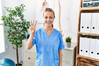 Ağrı kesici kliniğinde çalışan beyaz bir kadın üç numarayı gösterip işaret ederken kendinden emin ve mutlu bir şekilde gülümsüyor.. 