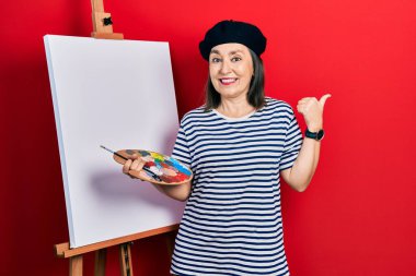 Orta yaşlı İspanyol kadın paletle resim çiziyor. Resim sehpası arkasını işaret ediyor. El ve baş parmakları havada, kendinden emin gülümsüyor. 