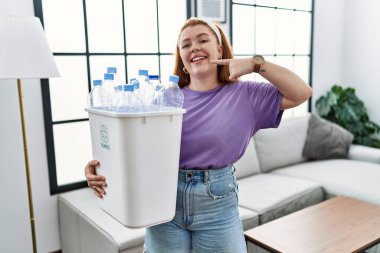 Geri dönüşüm sepetini elinde tutan, elinde plastik şişelerle gülümseyen ve parmaklarını ve ağzını gösteren kızıl saçlı genç bir kadın. diş sağlığı kavramı. 