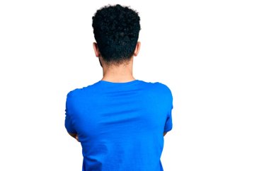 Sakallı, sıradan mavi tişörtlü genç bir Arap. Kollarını kavuşturup arkasını kolluyor. 