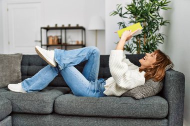 Evdeki kanepede uzanmış kitap okuyan beyaz bir kadın.