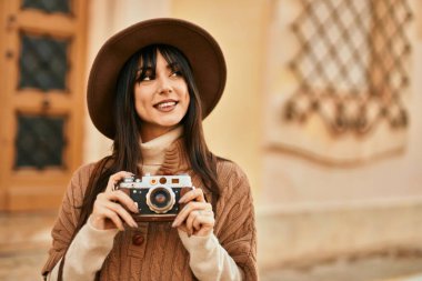Kışlık şapkalı esmer kadın şehrin dışında klasik kamera kullanarak gülümsüyor.