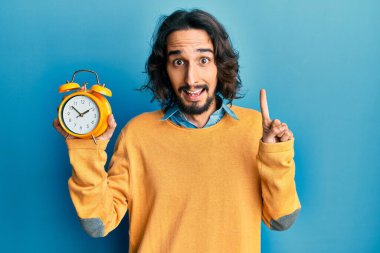 Çalar saati tutan genç İspanyol bir adam bir fikir ya da soruyla gülümsüyor ve mutlu bir yüzle işaret ediyor. 