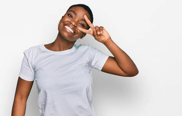 顔の上に指で平和のシンボルを行うカジュアルな白いTシャツを着て若いアフリカ系アメリカ人の女性 勝利を示す明るい笑顔 — ストック写真