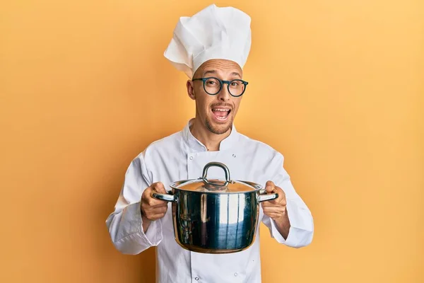 Bald man with beard wearing professional cook holding cooking pot celebrating crazy and amazed for success with open eyes screaming excited.