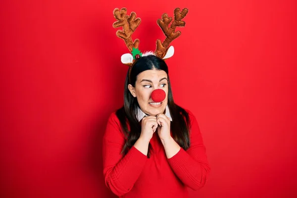 若いですヒスパニック女性身に着けています鹿クリスマスの帽子と赤い鼻笑い神経と興奮で手で顎見ますザ側 — ストック写真