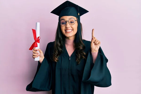 若いですヒスパニック系の女性を着て卒業制服を保持卒業証書笑顔でアイデアや質問を指差す指で幸せな顔 ナンバーワン — ストック写真