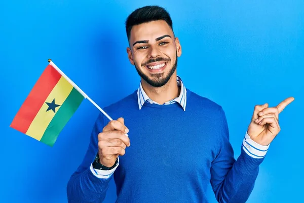 Joven Hispano Con Barba Sosteniendo Bandera Ghana Sonriendo Feliz Señalando —  Fotos de Stock