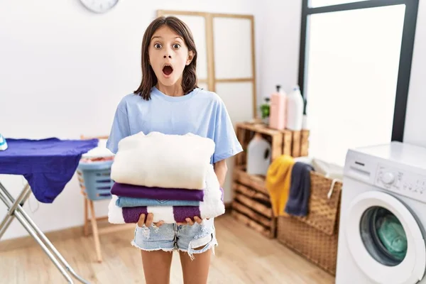 Young hispanic girl holding folded laundry afraid and shocked with surprise and amazed expression, fear and excited face.