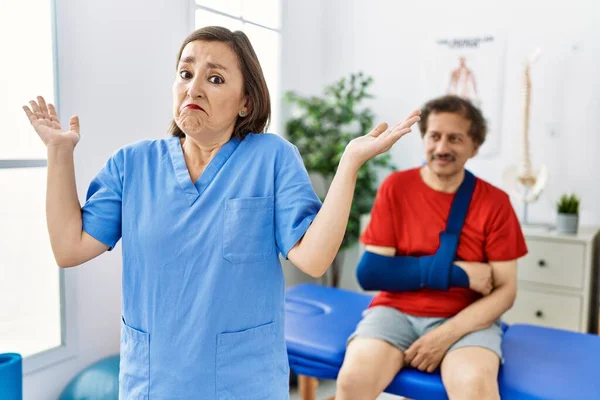 Middle age doctor woman with patient with arm injury at rehabilitation clinic shouting and screaming loud to side with hand on mouth. communication concept.