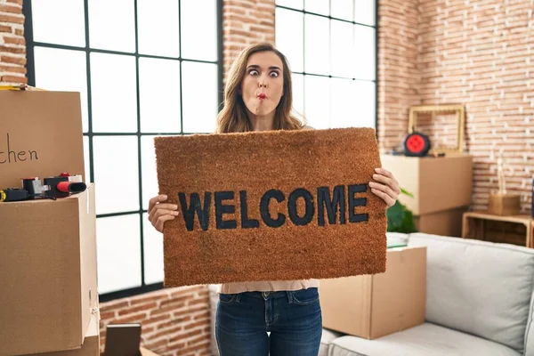 Young woman holding welcome doormat at new home making fish face with mouth and squinting eyes, crazy and comical.