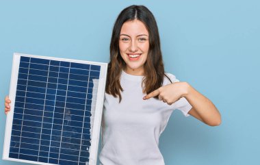 Fotovoltaik güneş panelini tutan genç ve güzel bir kız mutlu ve gururlu bir gülümsemeyi işaret ediyor. 