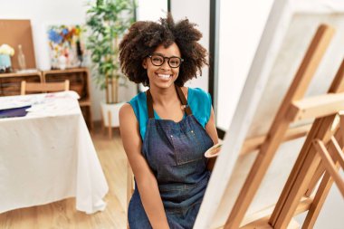 Güzel Afro saçlı Amerikalı kadın sanat stüdyosunda tuvali boyuyor. Pozitif ve mutlu görünüyor. Kendine güvenen bir gülümsemeyle dişlerini gösteriyor. 