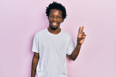 Genç Afro-Amerikan adam, günlük beyaz tişört giyer ve parmaklarıyla iki numarayı gösterirken kendinden emin ve mutlu gülümserdi.. 
