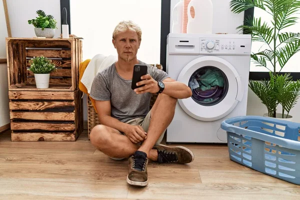 Genç Sarışın Adam Çamaşır Yıkıyor Akıllı Telefon Kullanıyor Şüpheci Gergin — Stok fotoğraf