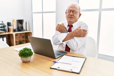 Ofiste çalışan kıdemli bir adam bilgisayarla kendine sarılıyor mutlu ve pozitif, kendinden emin gülümsüyor. Sevgi ve özgüven. 