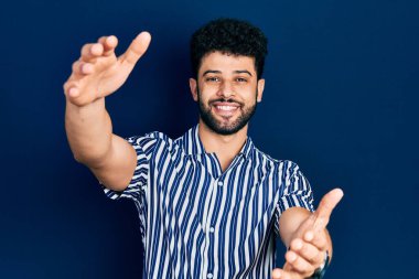 Sakallı, sıradan çizgili tişörtlü genç bir Arap kameraya sarılmak için gülümseyerek bakıyor. Mutluluğu kucaklayan neşeli bir ifade.. 