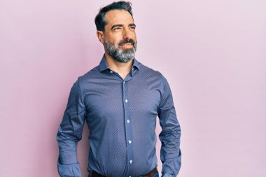 Sakallı, gri saçlı, iş kıyafetleri giyen, yüzünde gülümsemeyle yan yana bakan orta yaşlı bir adam. Kendinden emin gülen. 