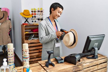 Young hispanic woman shopkeeper smiling confident scanning hat using bardcode reader at clothing store clipart