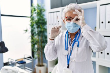 Gri saçlı, doktor üniforması giyen ve elinde şırıngayla gülümseyen yaşlı bir kadın. 
