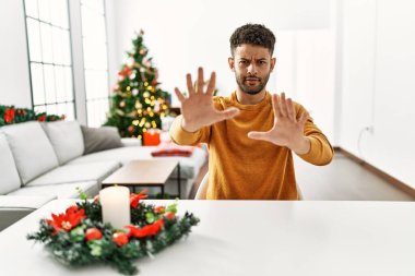 Arap genç adam yılbaşı ağacı tarafından masada oturuyor korku ve korku ifadesinden korkuyor jesti elleriyle durdurup şok içinde bağırıyor. panik konsepti. 